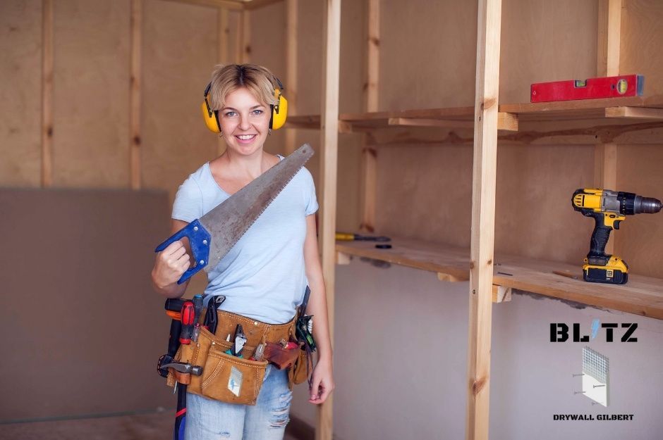 A drywall repair company worker wearing yellow safety ear plugs and a tool belt, holds a saw while working in a residential construction site with exposed wooden framing near W White Canyon Rd, and N Cherry Creek Rd in San Tan Valley, AZ.
