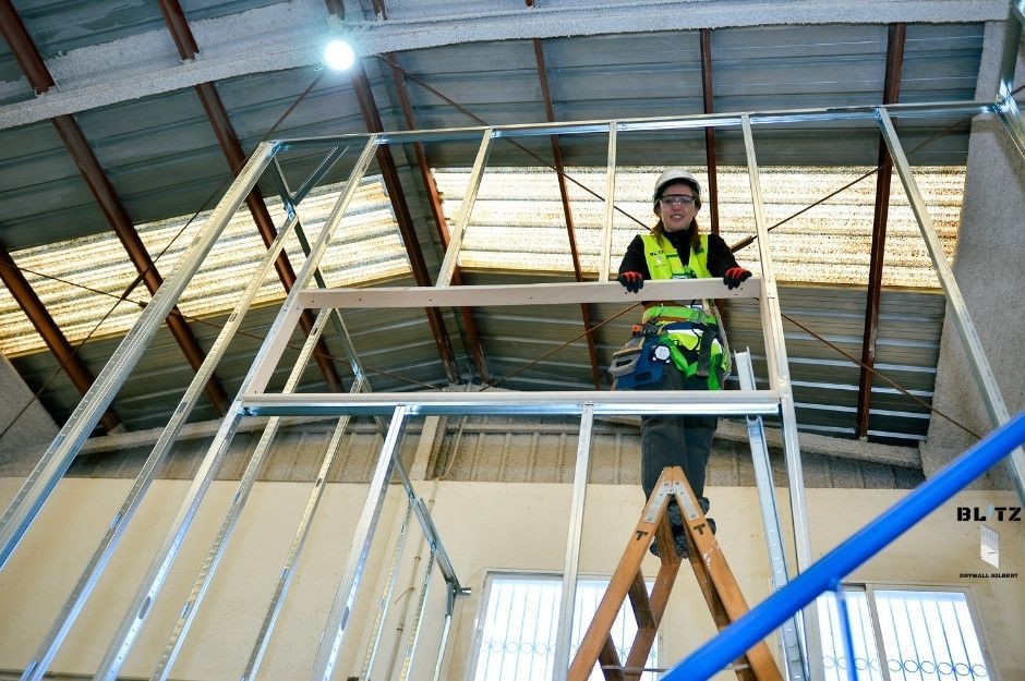 A drywall repair company worker in yellow high-visibility safety vest stands on metal scaffolding platform preparing to install or repair the drywall ceiling in large industrial warehouse with exposed steel beams. Located in Gainey Ranch, Scottsdale, AZ.