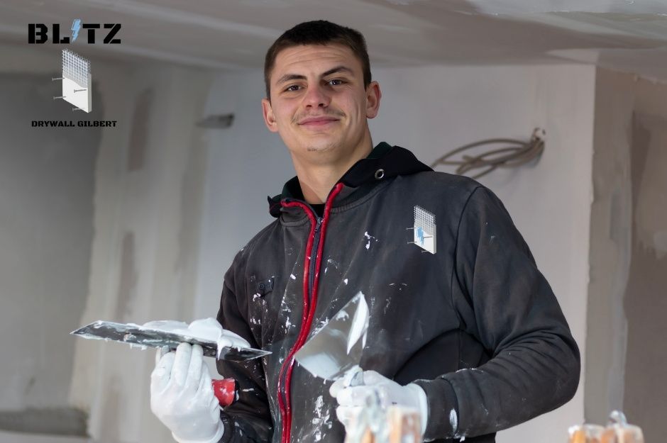 A drywall repair contractor in a dark hoodie stands in an unfinished interior space, holding a drywall tool. He is a skilled specialist working on a residential project near Candela Ln, and Dolce Vita Wy, Apache Junction, AZ.