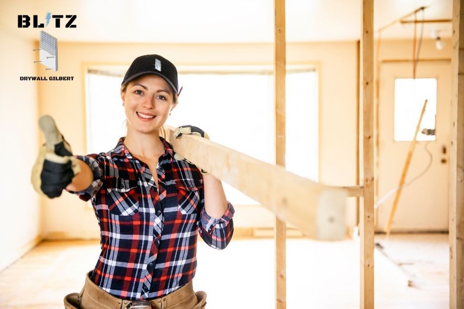A drywall repair contractor wearing a black cap and red plaid flannel holds one piece of wooden lumber in a bright residential construction site with exposed wood framing near Calliandra Ct, and S Superstition Mountain Dr in Gold Canyon, AZ.