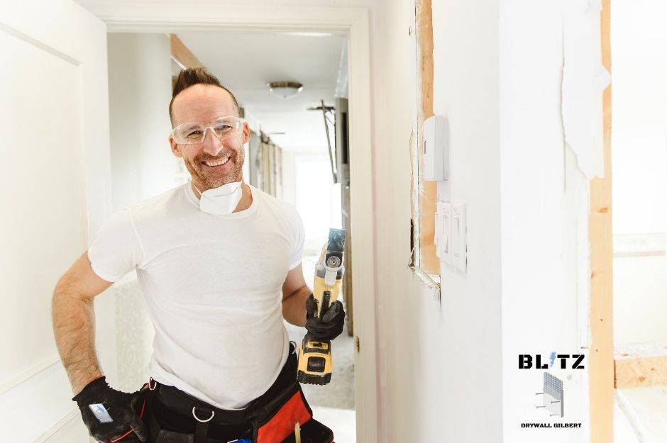 A drywall repair contractor wearing safety glasses and white shirt holds a reciprocating saw while working on a residential renovation project. He stands in a hallway with exposed framing and drywall dust on the floor near W Polk St, and N 166th Ln in Goodyear, AZ.