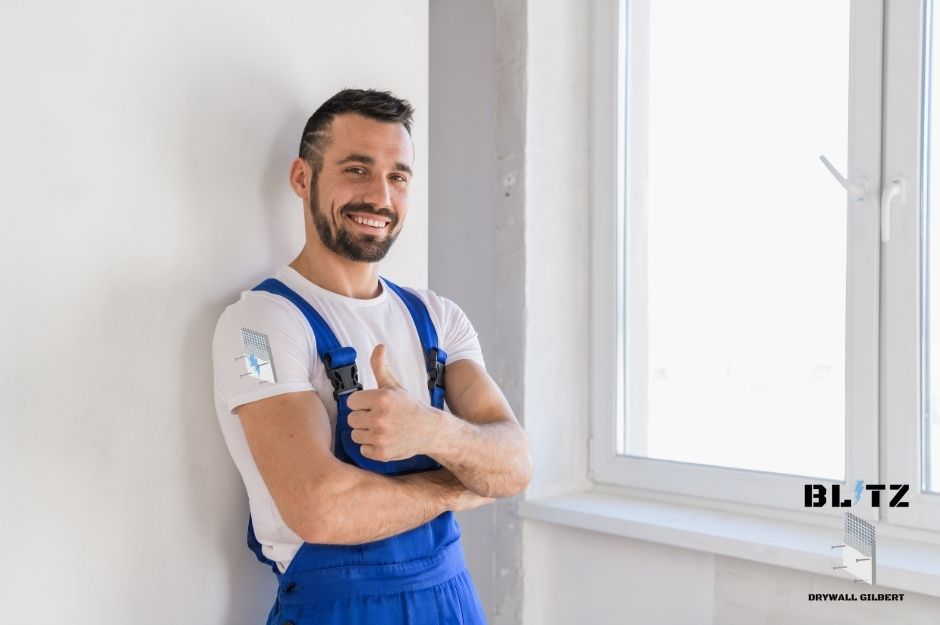 A drywall repair contractor in blue overalls and white t-shirt gives a thumbs up while standing against a white wall in a commercial building. He has his arms crossed and is smiling confidently in a clean, modern workspace near S Calle Vauo Nawi, and E Calle Sewa in Guadalupe, AZ.