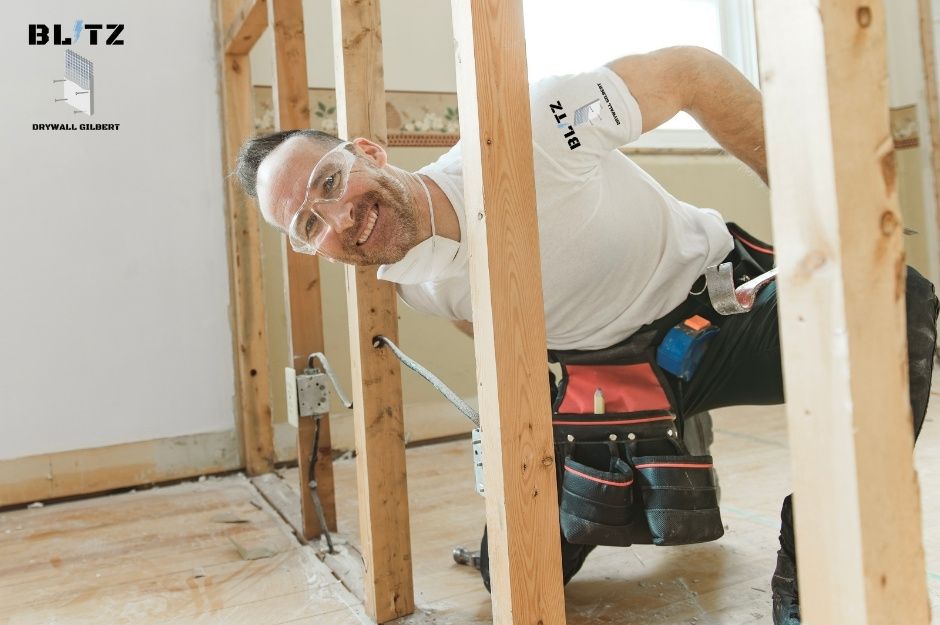 A drywall repair contractor wearing safety glasses and a white shirt. He smiles while looking over a wall framing in a residential project near 3rd St and Coronado St, Mammoth, AZ.