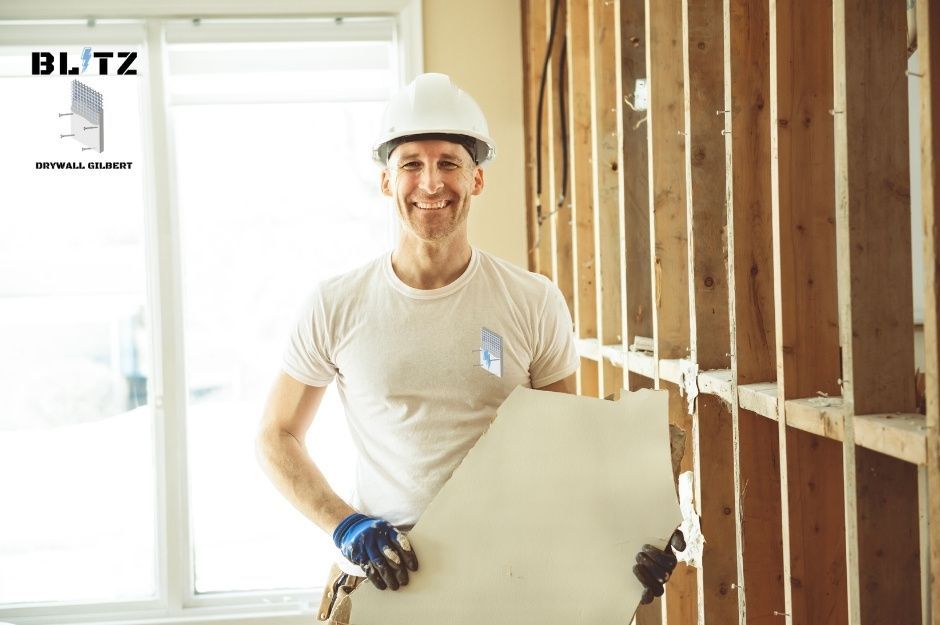 A drywall repair contractor in a white hard hat and t-shirt holds a drywall sheet while smiling in a residential construction site. He wears blue work gloves and stands among exposed wooden framing with natural light from windows. The renovation project is near N 90th Ln, and N 91st Ln in Peoria, AZ.