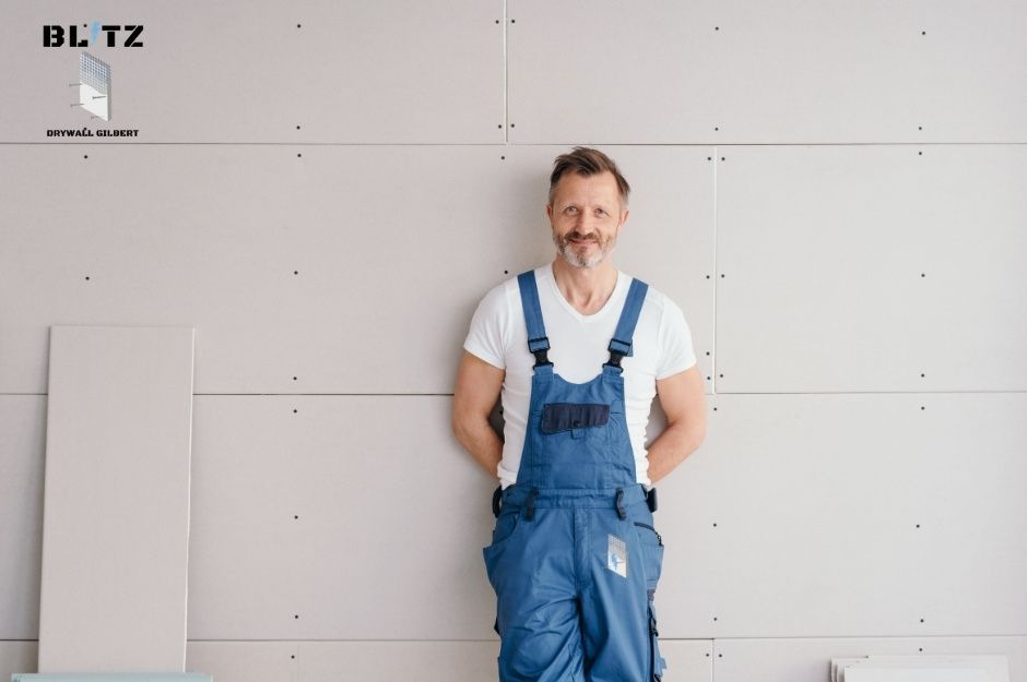 A drywall repair handyman in blue overalls and white t-shirt stands with his hands behind his back against freshly installed drywall in a commercial construction space. He is positioned next to drywall sheets with visible screw holes near W Crittenden Ln, and N 302nd Dr in Buckeye, AZ.