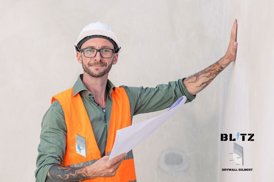A drywall repair handyman with glasses and white hard hat smiles while holding blueprints and leaning his hand against the white drywall inside a commercial construction site. He wears an orange safety vest over green shirt near W Denton Ave, and N 184th Dr, Citrus Park, AZ.