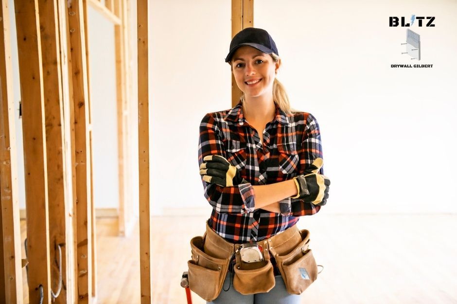 A drywall repair specialist in a plaid shirt and baseball cap stands with arms crossed in a residential construction site. She wears work gloves and a leather tool belt while positioned among exposed wooden framing near E Kino Rd, and E Santa Rita Rd in Cordes Lakes, AZ.