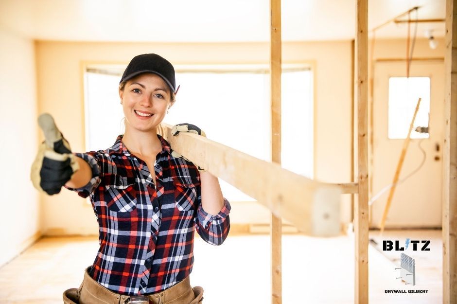 A drywall repair specialist in a plaid shirt and black cap carries lumber on her shoulder in a commercial construction site. The workspace shows ongoing commercial construction and framing work near S Burton Rd, and High Rd in Spring Valley, AZ.