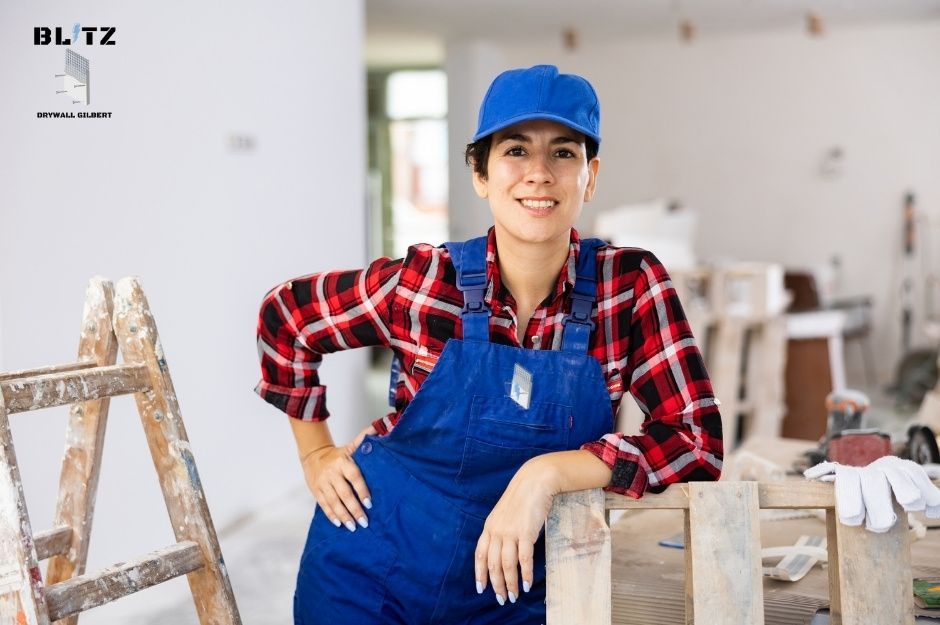 A drywall repair technician wearing blue overalls, red plaid shirt, and blue cap, smiling while leaning against wooden construction materials. Female worker appears relaxed in interior residential construction space with wooden ladder visible in background near E Villa Cassandra Way, and N 36th Pl, Cave Creek, AZ.
