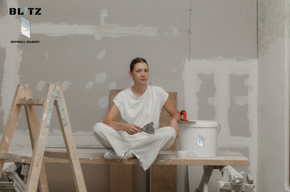 A drywall repair technician in white work clothes sits on a wooden workbench holding a putty knife in a commercial renovation space. She's positioned next to a paint bucket with a step ladder nearby, surrounded by patched drywall walls near W Willow Ave, and N 125th Ave in El Mirage, AZ.