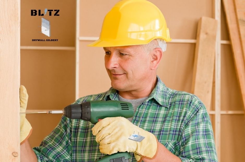 A drywall repair technician in a yellow hard hat and green plaid shirt uses a power drill on a residential construction project. He wears work gloves while operating the tool in a renovation with exposed wooden framing and drywall materials visible in the background near N Sapphire Ave, and W Steen Rd in Maricopa, AZ.