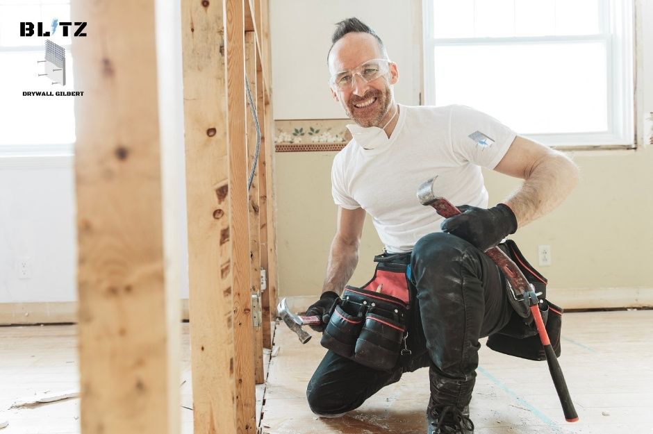 A drywall repair technician wearing safety glasses and black gloves holds a hammer while kneeling in a residential renovation space. He's dressed in a white shirt with a tool belt, working on a home construction project with exposed wood framing and natural lighting near W Surrey Dr, and N 141st LN in Surprise, AZ.