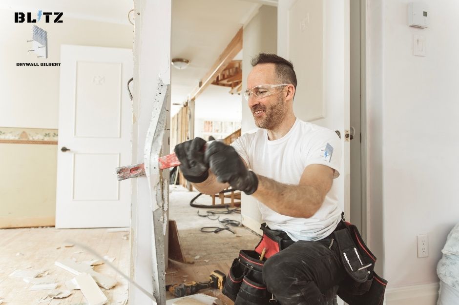 A drywall repair technician wearing safety glasses and black gloves uses a crowbar on a residential renovation project. He's dressed in a white shirt with a tool bag, working on drywall in a home under construction near N Christa Way, and W Irene Ln in Tolleson, AZ.