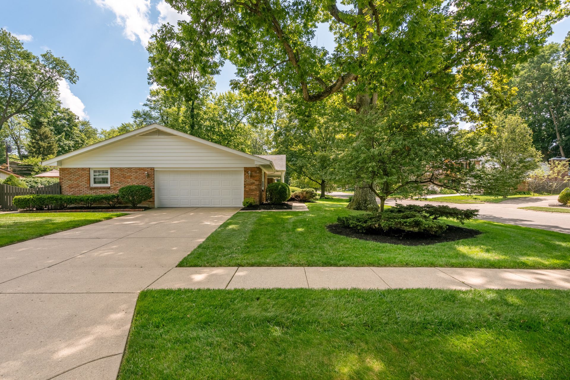 Brick house with garage, driveway, and lush green lawn, under blue sky with trees.