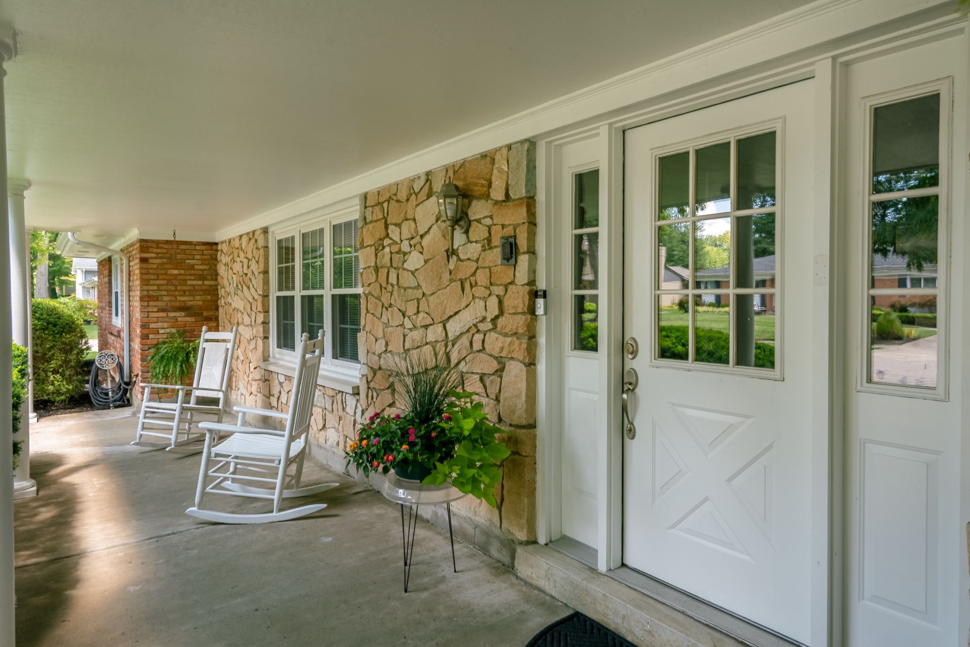 Covered porch of a house with white double doors, rocking chairs, stone wall, and potted plants.