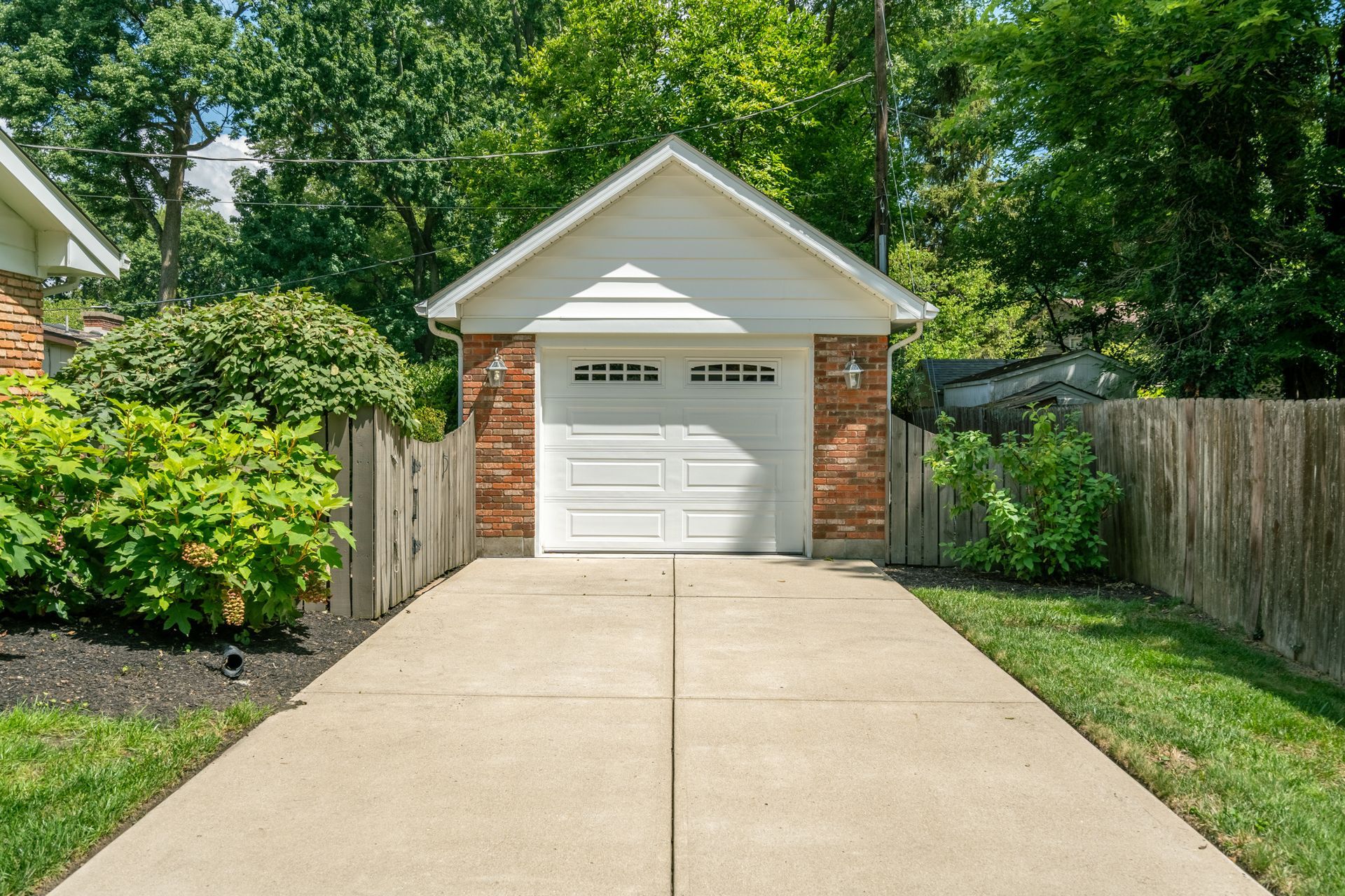Concrete driveway leads to a white garage with brick accents, surrounded by greenery and a wooden fence.