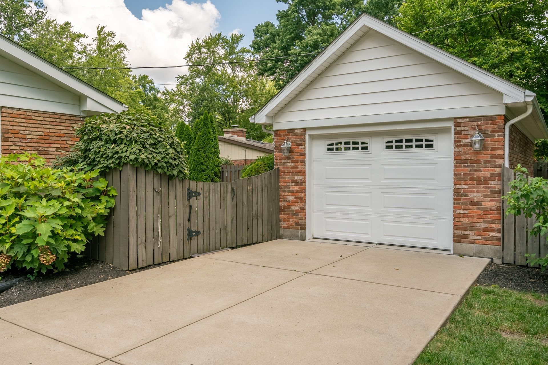 Garage with white door, brick walls, and concrete driveway. Wooden fence and greenery flank the driveway.