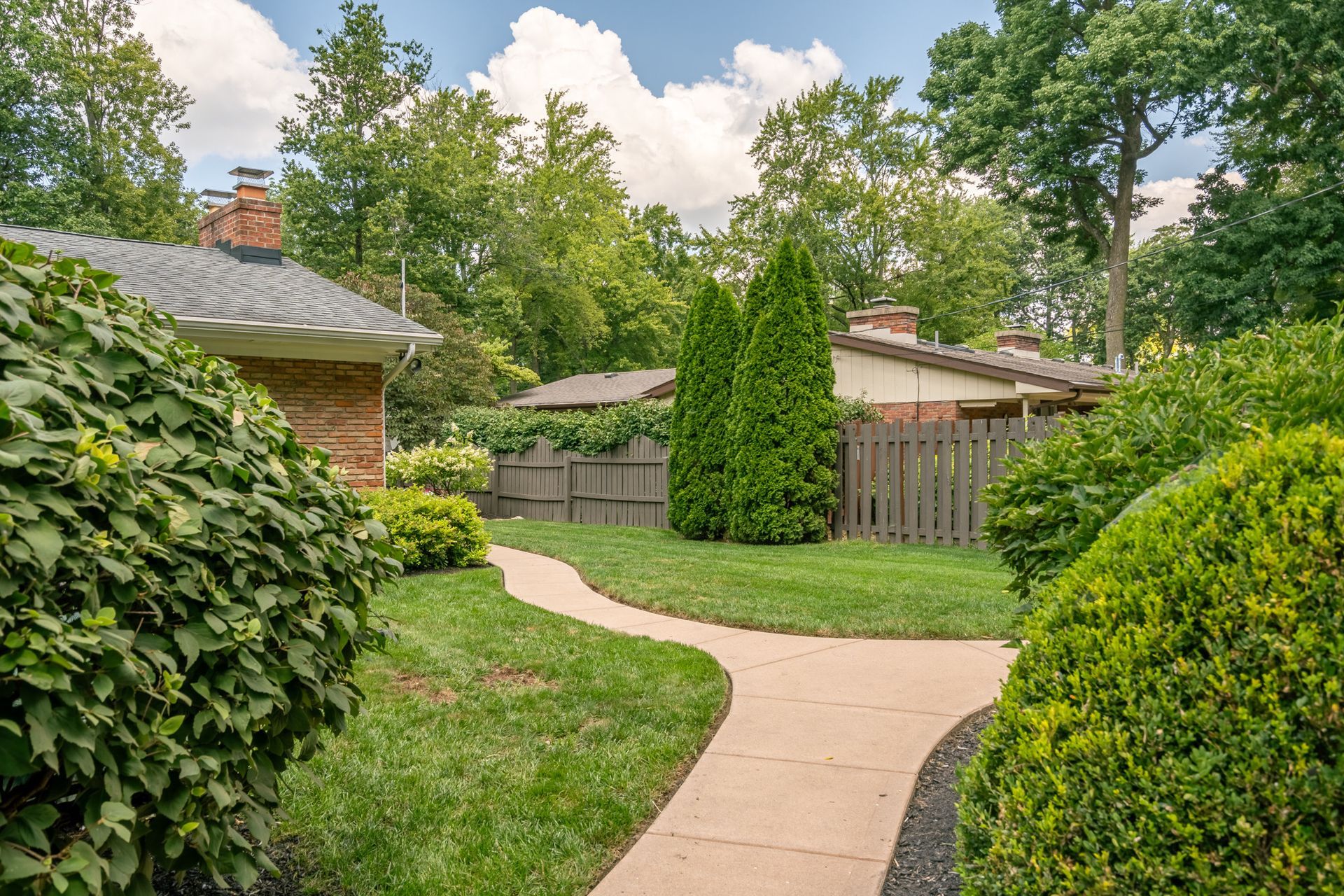 Pathway through a yard with green bushes and trees, leading toward houses with fences.
