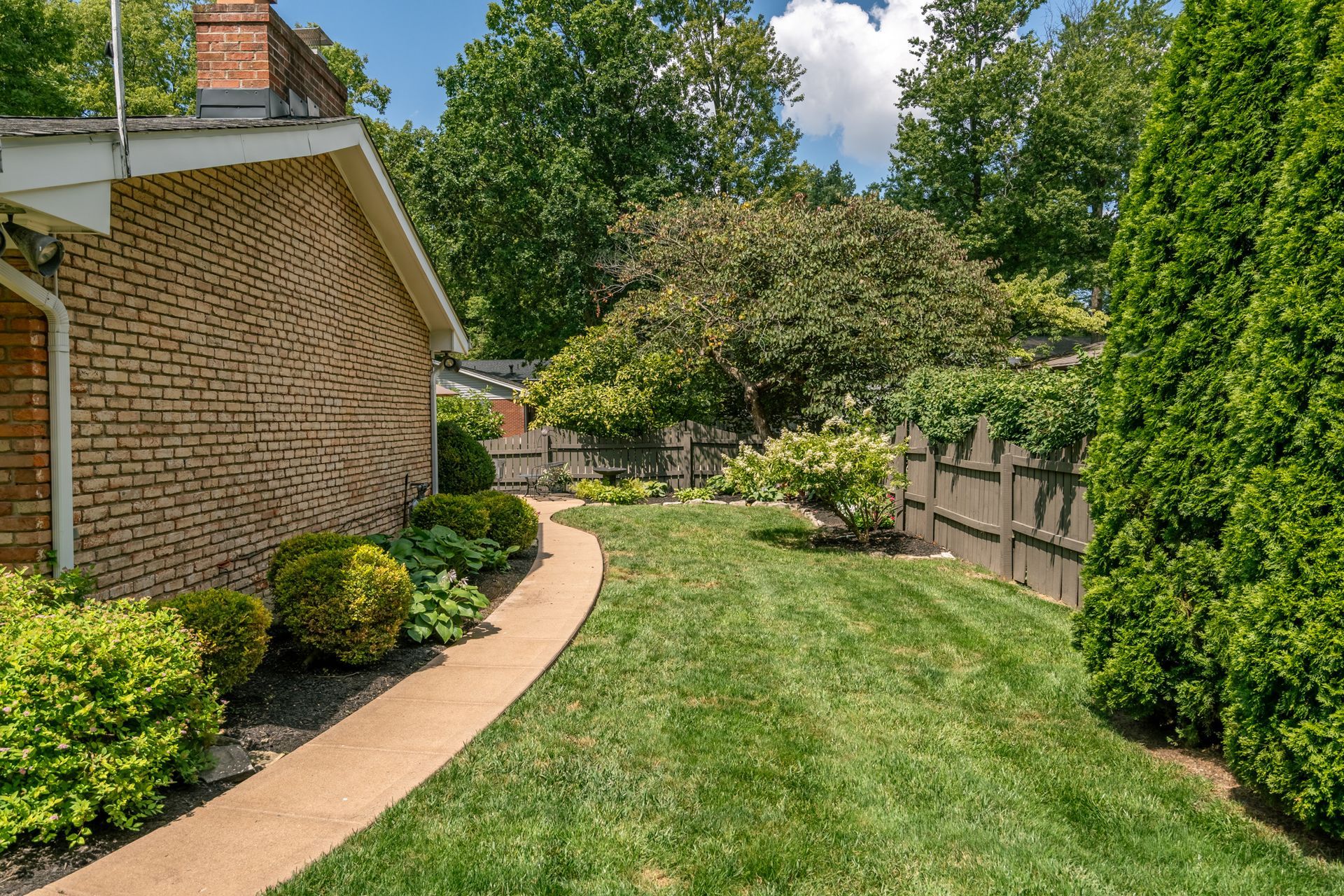 Brick house exterior with a curved walkway, green lawn, and shrubbery. Wooden fence and trees in background.