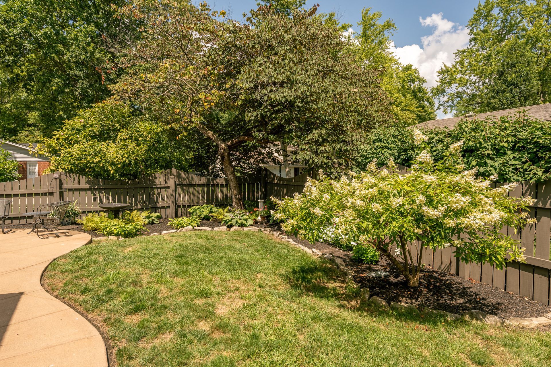 Lush green backyard with trees, shrubs, and a curved stone pathway, enclosed by a wooden fence.