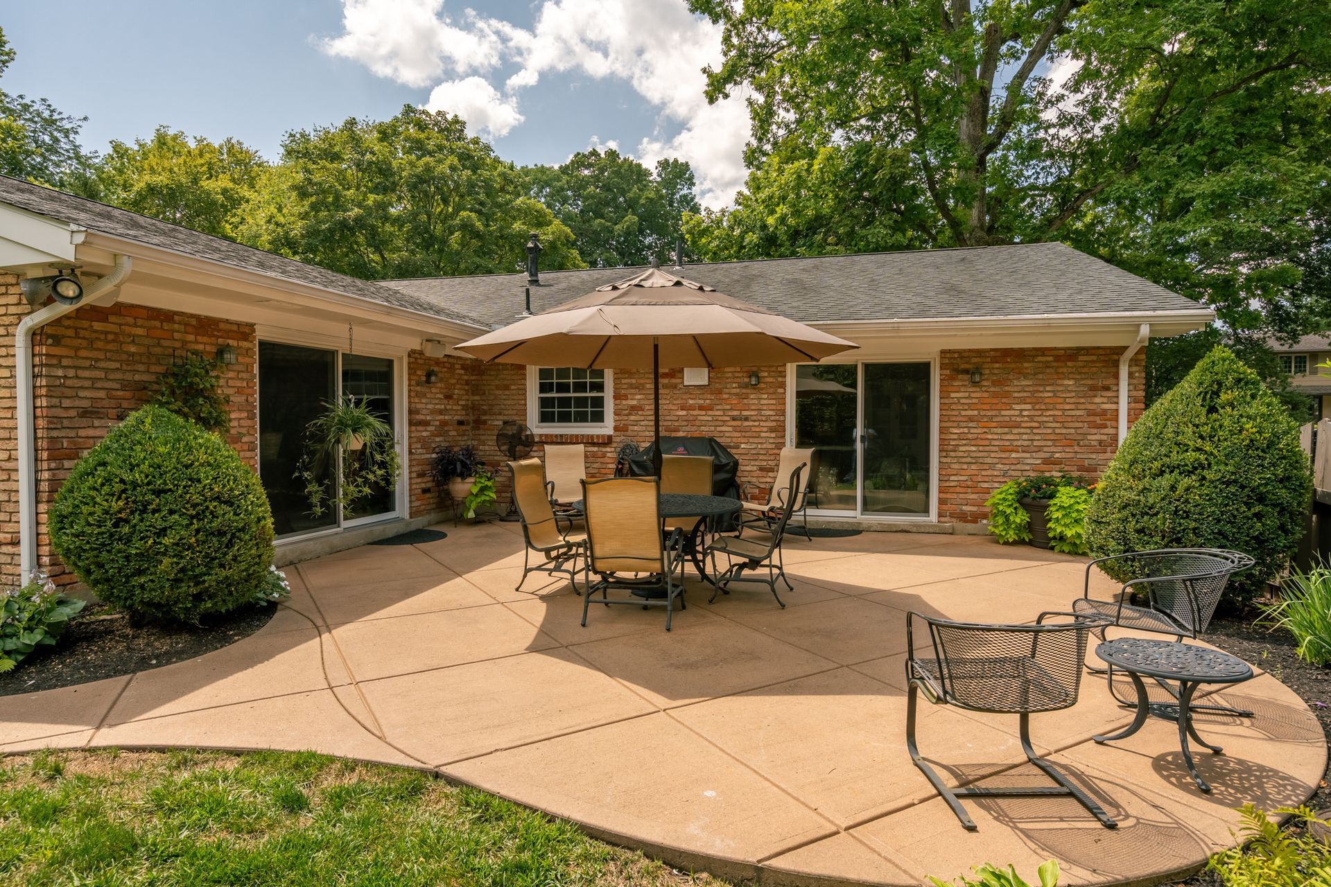 Patio with outdoor furniture, umbrella, and a brick house. Green trees and grass in the background.