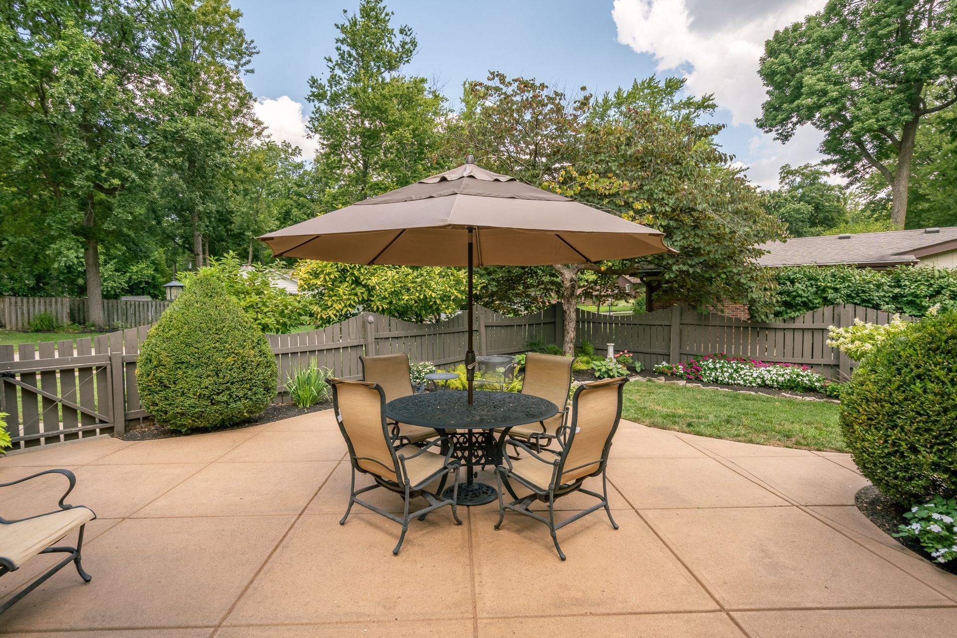 Patio with table, chairs, and umbrella in a backyard with trees and a fence.
