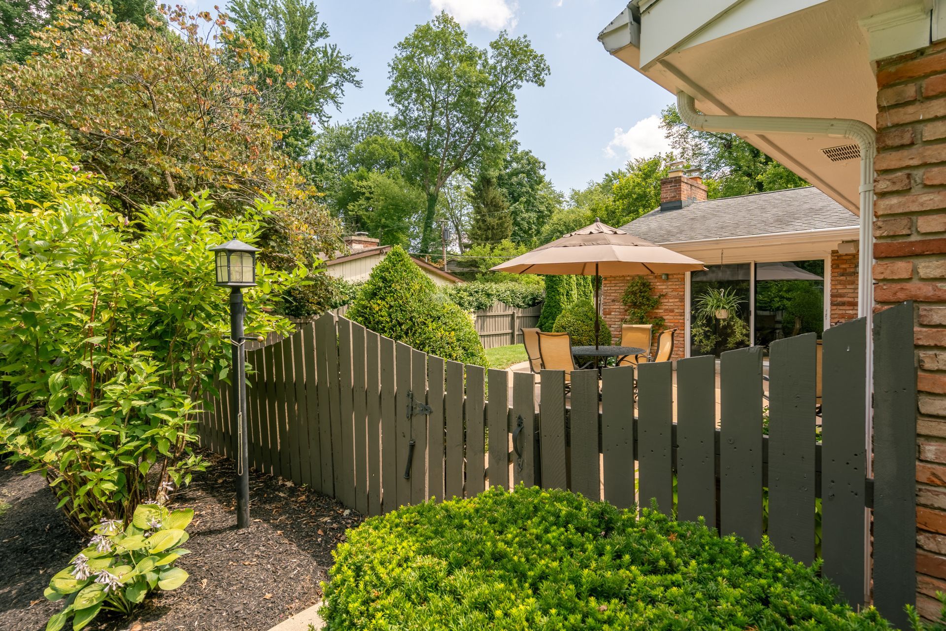 Gray picket fence with lush landscaping in front of a patio with an umbrella.