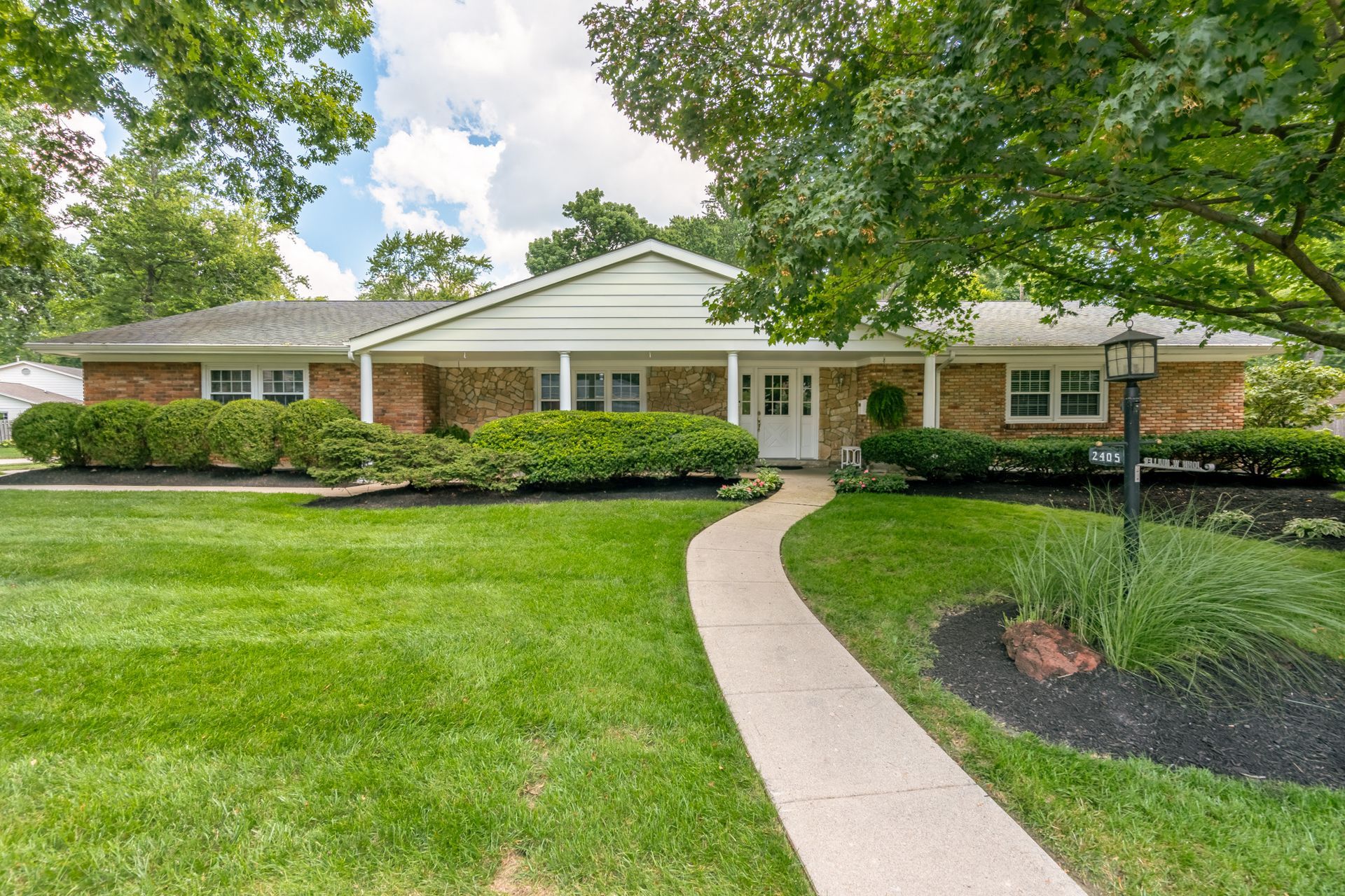 Brick ranch-style house with a pathway through a well-maintained lawn, framed by bushes and trees.