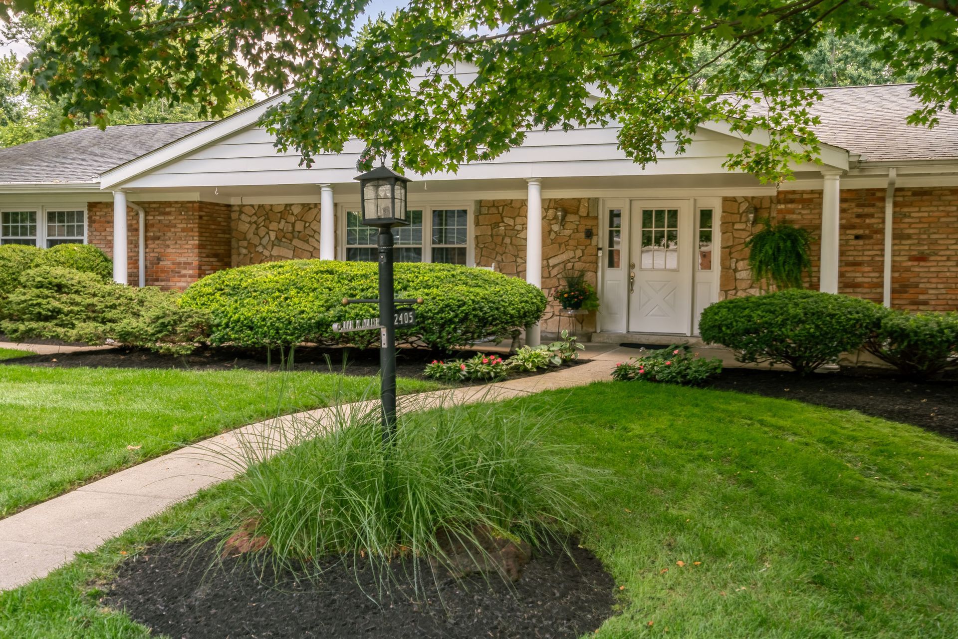 Ranch-style house with brick facade, white trim, and a walkway leading to the front door. Lush green lawn and shrubs.