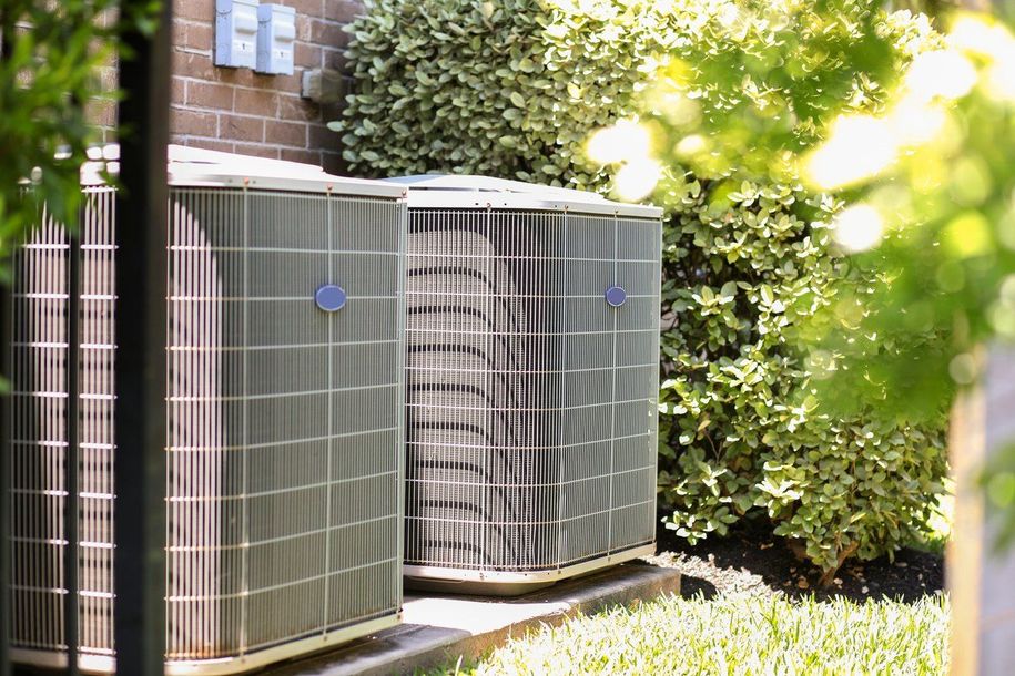 Two outdoor air conditioning units next to a brick building and surrounded by greenery.
