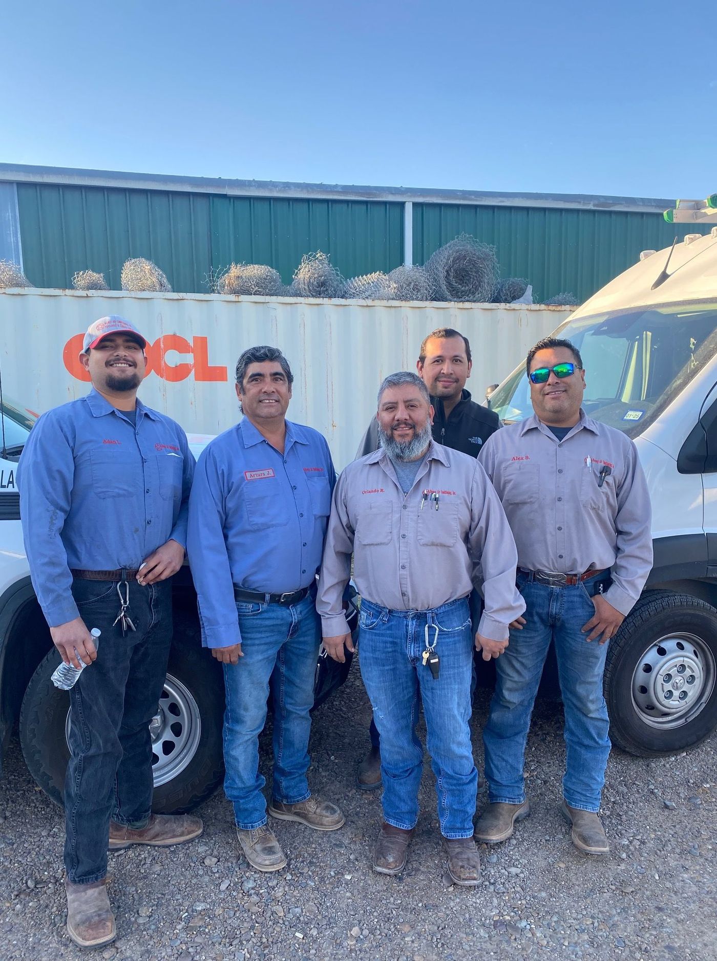 Five men in work uniforms standing next to a white van and container; outdoors.