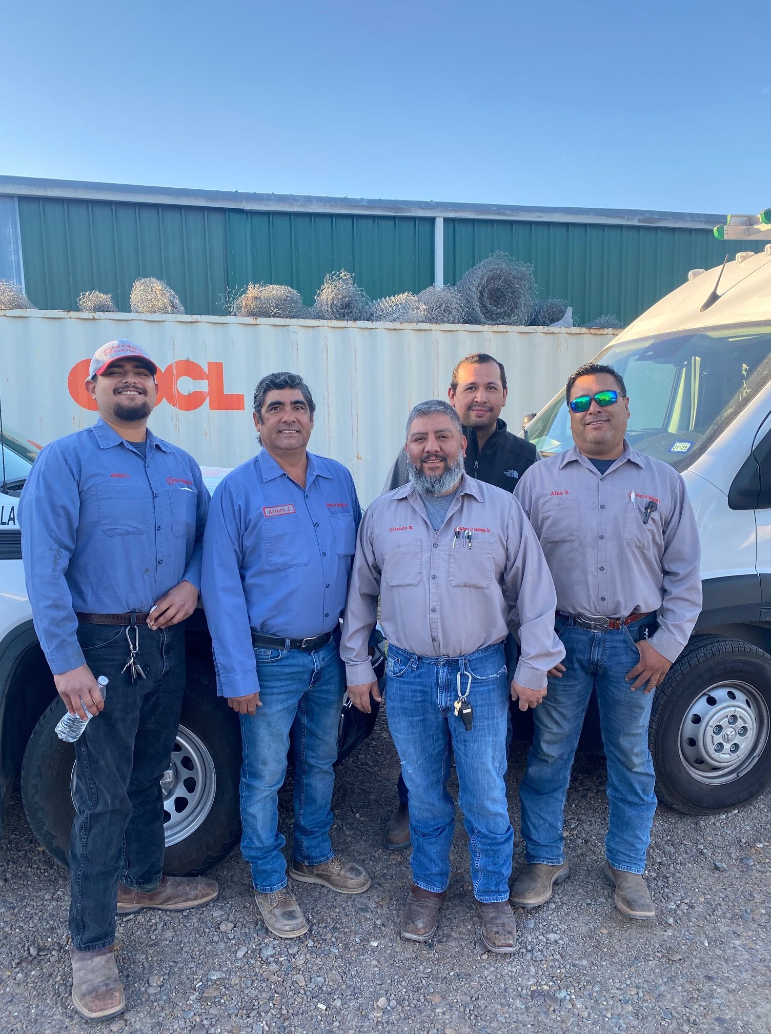 Five men in work uniforms standing next to a white van and container; outdoors.