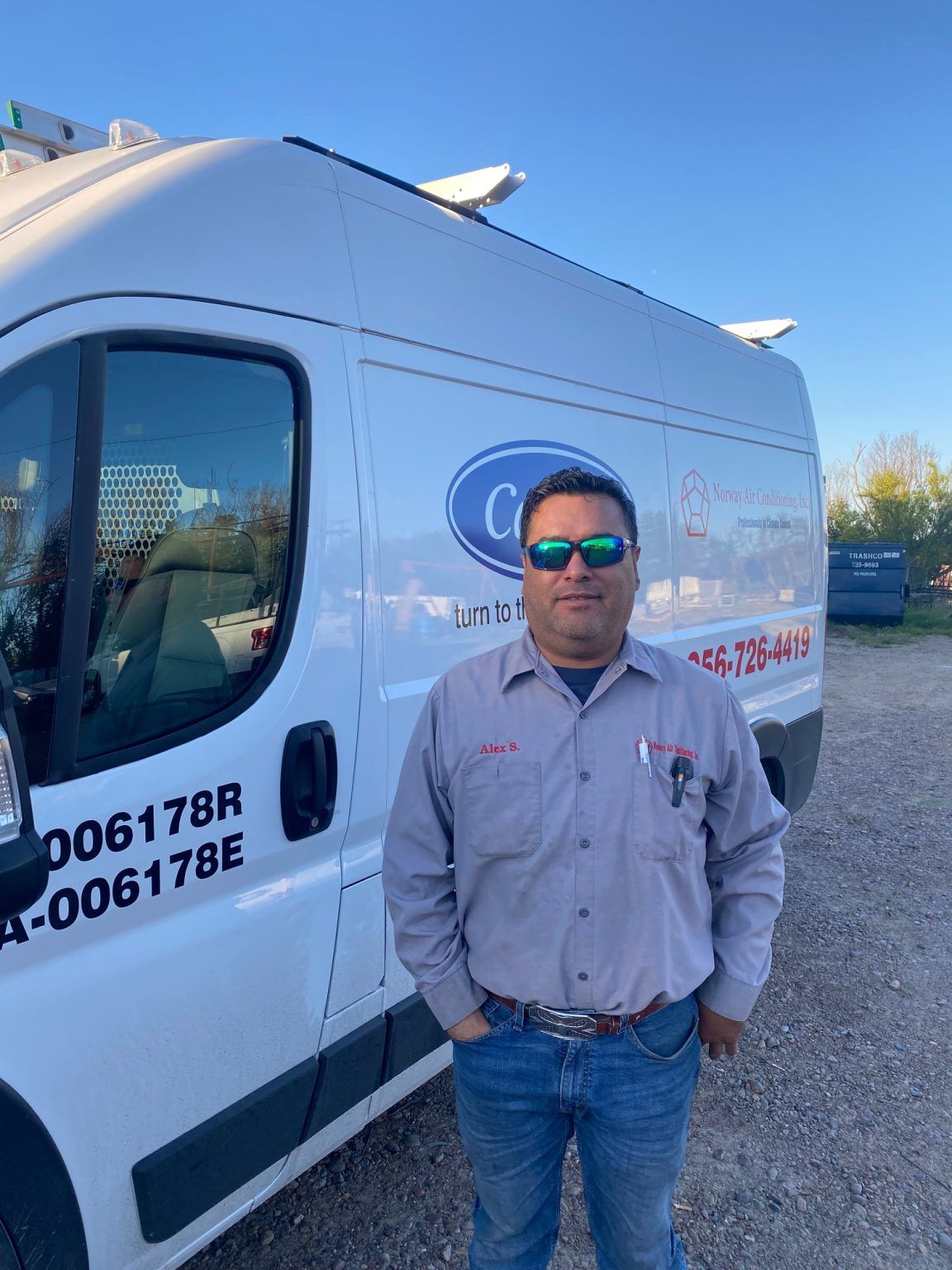 Man in work uniform stands in front of a white company van, hands in pockets, outdoors.