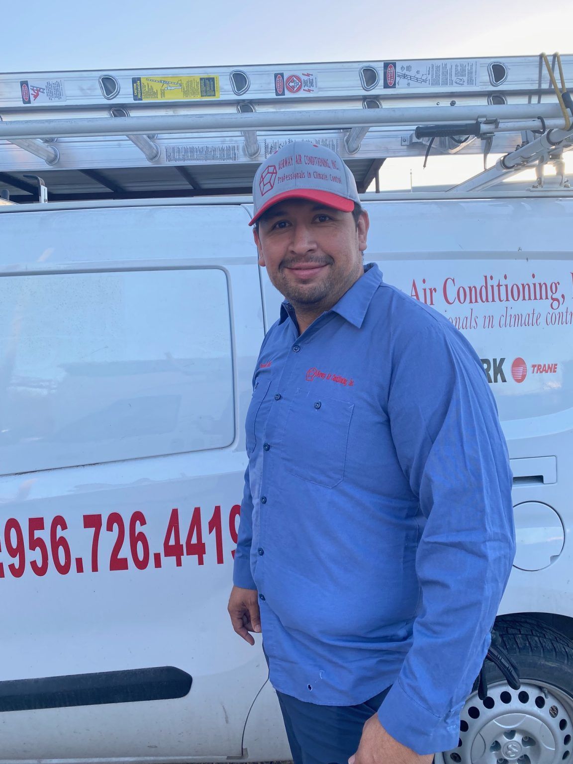 Man in work uniform smiles in front of a white van with phone number on the side.