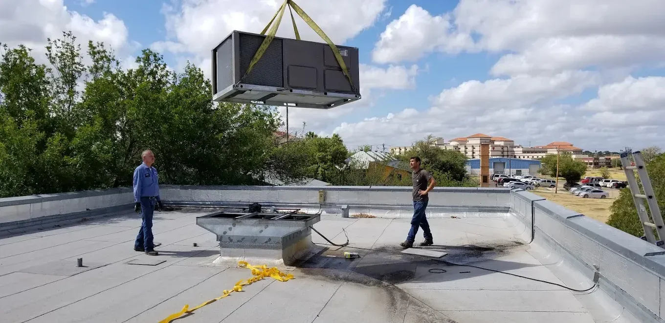 HVAC unit being lowered onto a roof by a crane. Two men are standing nearby on the roof.