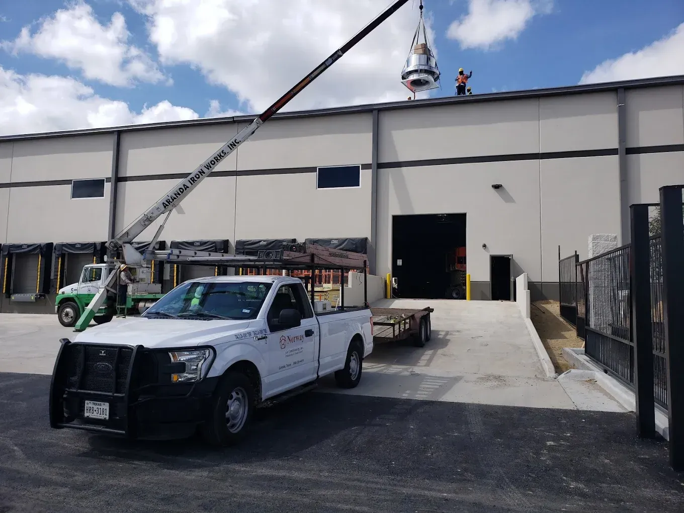 White truck and crane lifting a silver object onto the roof of a warehouse. Two people on roof.
