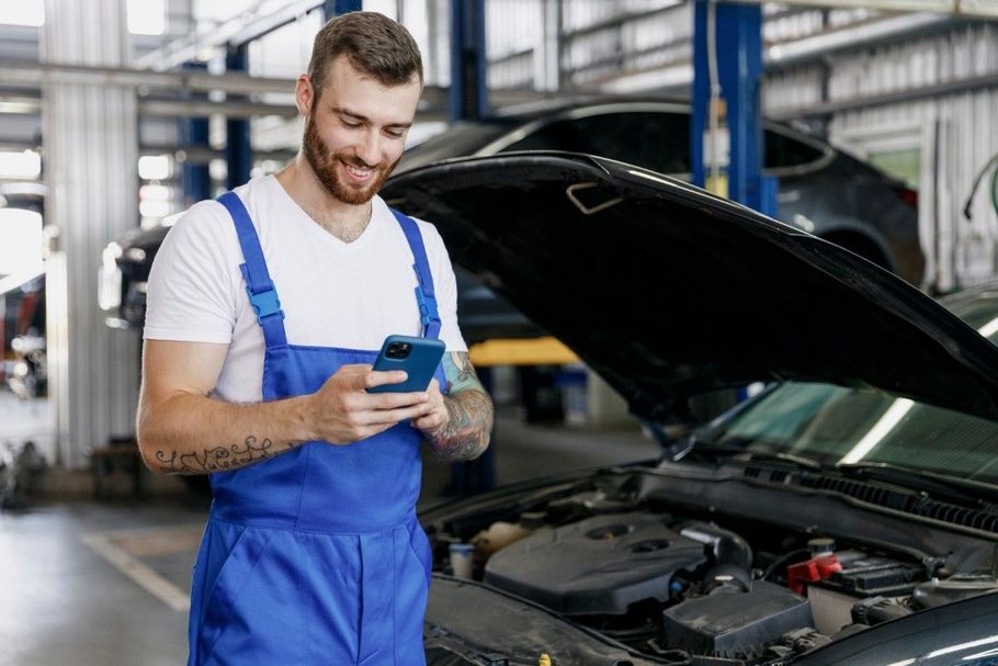 Mechanic in blue overalls checking a car engine with a handheld device in a garage