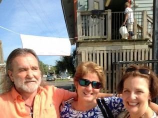 Three people smiling in front of a building; a person on the balcony. Bright sunny day, white cloth.