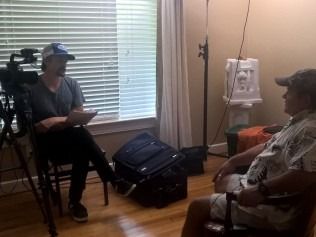 Two men in a room, one interviewing the other. A camera and luggage are nearby.