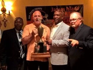 Men holding trophy in front of a banner. One in orange shirt, others in suits. James Island banner.