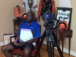 Man seated for interview, camera on tripod. Brown, orange decor.