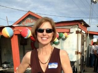 Woman in sunglasses smiles, in front of a colorful shack with beach balls, 