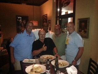 Five men at a restaurant table, smiling. One gives a thumbs-up. Plates of food are present.
