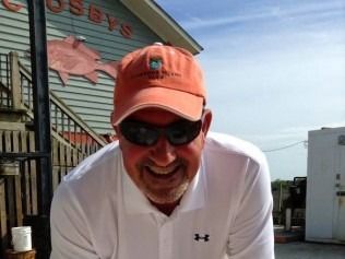 Man in orange cap and sunglasses smiles outdoors near a building with a fish sign.