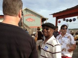 People gathered outside a restaurant, Crabby's, with a red fish sign. One man in a cap smiles.