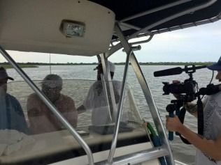 Boat with people filming; water and overcast sky in the background. A person holding a camera is visible.