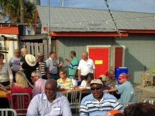 Group of people at an outdoor gathering, seated at tables and near a building with a red door.