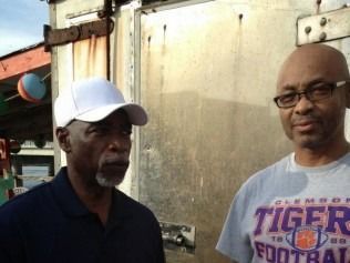 Two men stand outdoors; one wears a white cap, the other a Clemson Tigers t-shirt.
