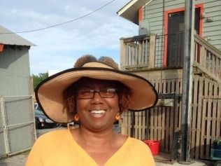 Woman wearing a wide-brimmed hat smiles outdoors, background of weathered buildings, orange top, glasses.