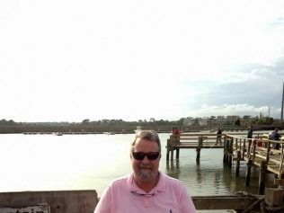 Man in sunglasses and pink shirt in front of a body of water and a pier.